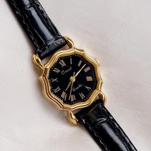 May include: A gold-toned Orient Quartz watch with a black face and Roman numeral hour markers. The watch has a black leather strap. The watch face is octagonal in shape.
