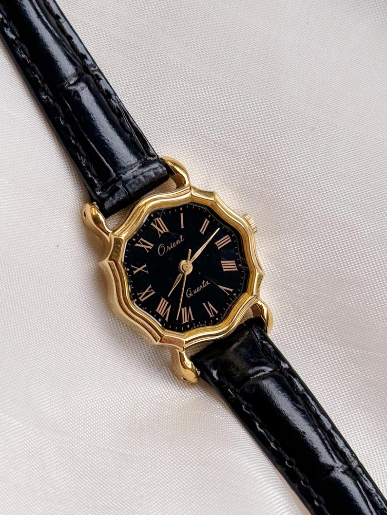 May include: A gold-toned Orient Quartz watch with a black face and Roman numeral hour markers. The watch has a black leather strap. The watch face is octagonal in shape.