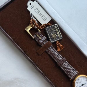 May include: A gold-tone wristwatch with a white dial, black Roman numerals, and a brown textured leather strap. The watch is inside a brown velvet-lined box with a white tag labeled G 28869 and a rectangular tag reading Longines 150.