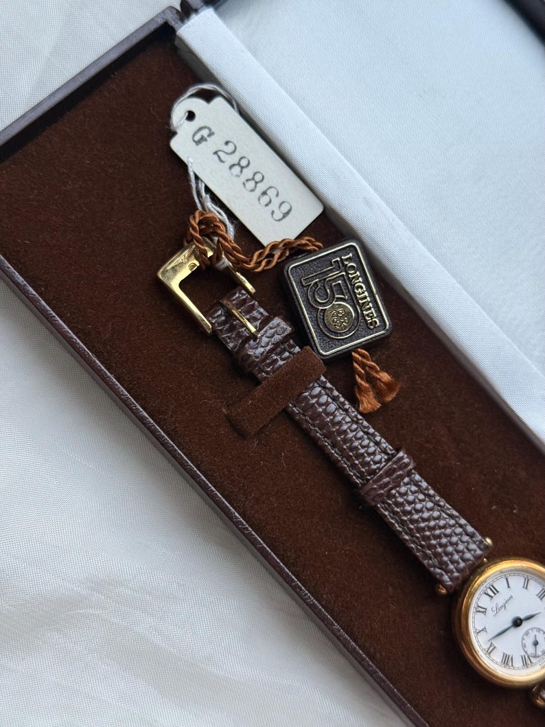 May include: A gold-tone wristwatch with a white dial, black Roman numerals, and a brown textured leather strap. The watch is inside a brown velvet-lined box with a white tag labeled G 28869 and a rectangular tag reading Longines 150.