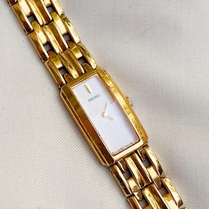 May include: Gold Seiko watch with a rectangular white face and gold hands. The watch band is made of interlocking gold links. The watch face has the word "SEIKO" printed on it. A classic timepiece.