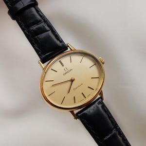 Pode incluir: Um relógio Omega Genève dourado com uma pulseira de couro preta. O mostrador do relógio é de cor ouro claro com marcadores e ponteiros pretos. A pulseira é texturizada e o relógio está sobre um fundo branco.