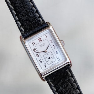 Puede incluir: Un reloj Seiko Quartz rectangular con esfera blanca, números negros y una pequeña subesfera. El reloj tiene una caja plateada y una correa de cuero negro con costuras blancas. La esfera muestra los números del 1 al 12.