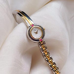 Puede incluir: Un reloj de pulsera bicolor con una correa de eslabones de metal plateado y dorado y una esfera redonda blanca con el texto TISSE. La caja del reloj está decorada con dos pequeñas piedras rosas en la parte superior e inferior y tiene manecillas doradas.