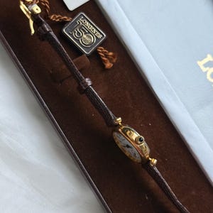 May include: A gold-tone Longines wristwatch with a thin brown textured leather strap. The watch features a round case with a small black crown. A metal tag attached to the strap displays the text Longines 150, and a white paper tag shows 28869.
