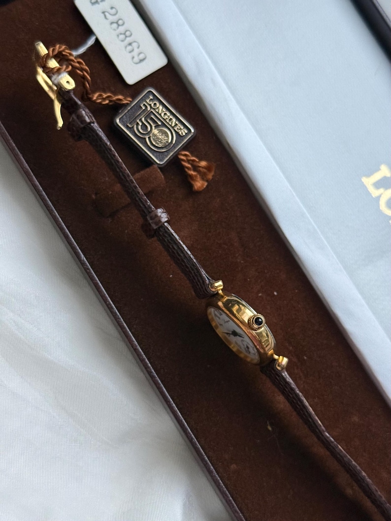 May include: A gold-tone Longines wristwatch with a thin brown textured leather strap. The watch features a round case with a small black crown. A metal tag attached to the strap displays the text Longines 150, and a white paper tag shows 28869.
