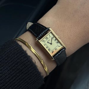 May include: A gold rectangular wristwatch with a cream-colored face and black Roman numerals. The watch has a black leather band and is paired with a gold bracelet. The watch face reads "Hermes Quartz".
