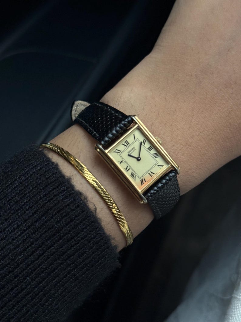 May include: A gold rectangular wristwatch with a cream-colored face and black Roman numerals. The watch has a black leather band and is paired with a gold bracelet. The watch face reads "Hermes Quartz".