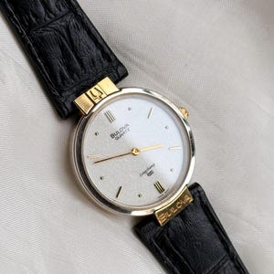 May include: A Bulova Quartz Longchamp watch with a silver face, gold hands, and a black leather band. The watch has gold accents and the Bulova logo is visible on the face and band.