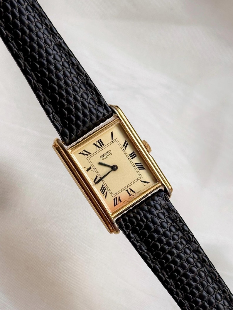 May include: A rectangular gold-toned Seiko Quartz watch with a cream-colored face and black Roman numerals. The watch has a black leather band. The watch face displays the Seiko Quartz logo. The watch is a classic style.