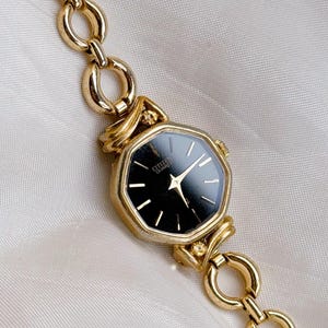Peut inclure: Une montre Citizen Quartz dorée avec un cadran noir et des aiguilles dorées. La montre a un cadran géométrique et est attachée à un bracelet à maillons dorés. La montre est présentée sur une surface blanche texturée.