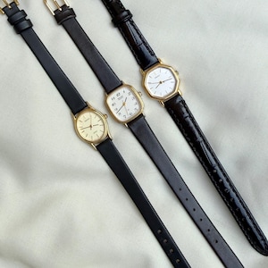 May include: Three vintage gold-toned watches with black leather bands. The watch faces are various shapes, including oval and rectangular. The watch bands are thin and have gold-colored buckles. The watches are displayed on a white surface.