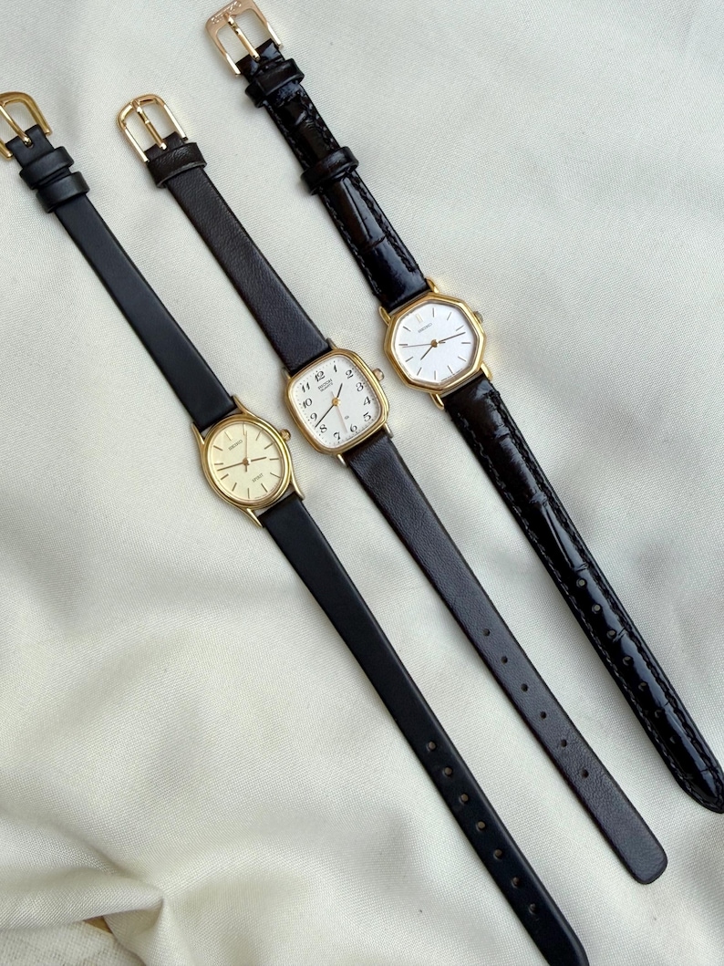 May include: Three vintage gold-toned watches with black leather bands. The watch faces are various shapes, including oval and rectangular. The watch bands are thin and have gold-colored buckles. The watches are displayed on a white surface.