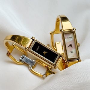 Puede incluir: Dos relojes Gucci dorados con esferas rectangulares descansan sobre una tela blanca. Uno tiene esfera negra, el otro esfera blanca nacarada. Ambos relojes tienen correas doradas. Se exhiben sobre una superficie blanca texturizada.