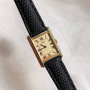 May include: A rectangular gold-toned Seiko Quartz watch with a cream-colored face and black Roman numerals. The watch has a black leather band with a textured pattern. The watch is a classic design.