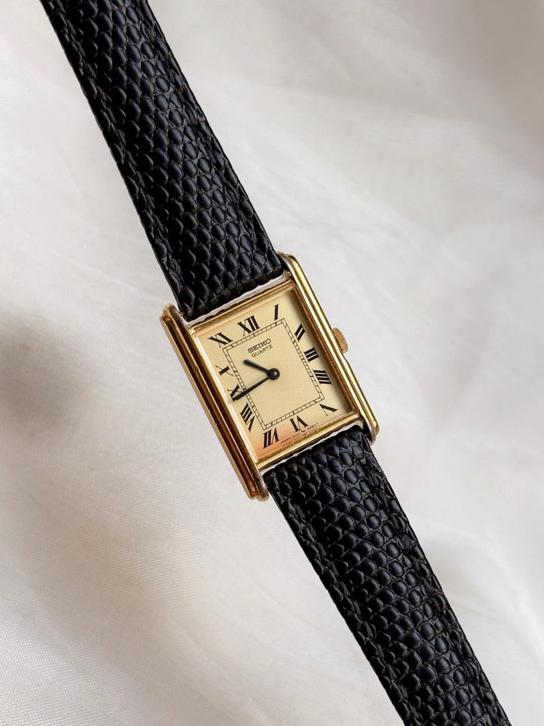May include: A rectangular gold-toned Seiko Quartz watch with a cream-colored face and black Roman numerals. The watch has a black leather band with a textured pattern. The watch is a classic design.