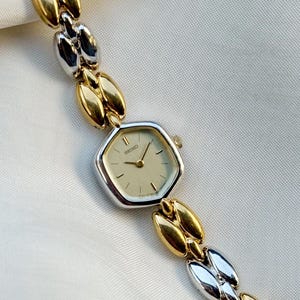 Könnte beinhalten: Eine zweifarbige Seiko-Uhr mit einem silber- und goldfarbenen Armband. Das Zifferblatt der Uhr ist cremefarben mit goldenen Zeigern und Markierungen. Das Armband besteht aus abwechselnd goldenen und silbernen Gliedern.
