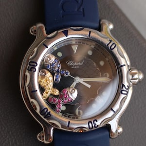 Könnte beinhalten: Eine luxuriöse Armbanduhr mit dunklem Zifferblatt und blauem Kautschukarmband. Die Uhr hat drei mit Juwelen besetzte Fische in Gelb, Blau und Rosa. Das Zifferblatt hat silberne Ziffern und Zeiger. Der Markenname "Chopard" ist sichtbar.
