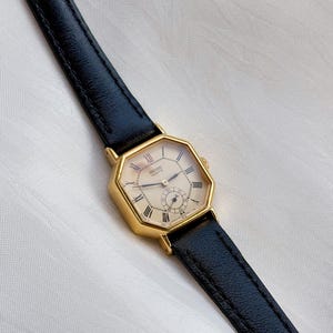 May include: A gold-toned Seiko Quartz watch with a cream-colored face and black leather band. The watch has Roman numeral hour markers and a small seconds dial. The watch case is an octagonal shape.