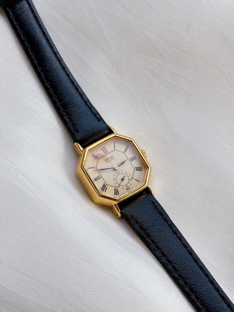 May include: A gold-toned Seiko Quartz watch with a cream-colored face and black leather band. The watch has Roman numeral hour markers and a small seconds dial. The watch case is an octagonal shape.
