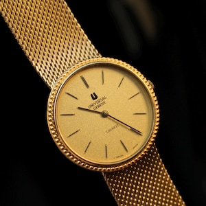 May include: A gold wristwatch with a mesh strap. The watch face is circular with a gold finish, displaying black hour markers and hands. The watch is marked "UNIVERSAL GENEVE QUARTZ" and "SWISS MADE".