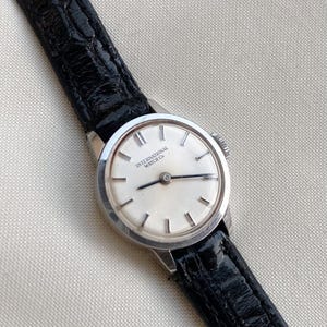 May include: A vintage wristwatch with a silver-toned case and a white face. The watch features black hands and hour markers. The black leather band is textured. The watch face reads "INTERNATIONAL WATCH Co."