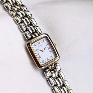 Puede incluir: Un reloj Seiko rectangular con esfera blanca, detalles dorados y números romanos. El reloj tiene una correa de eslabones plateada y dorada. El reloj se muestra sobre una superficie blanca.