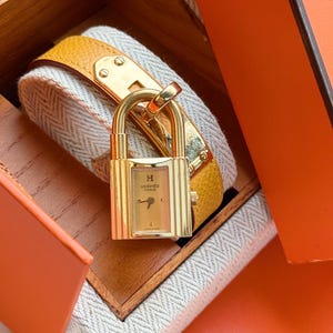 May include: A gold-toned watch with a rectangular face and a yellow leather strap. The watch face features the brand's logo and a small dial. The watch is secured with a padlock-style clasp. The watch is presented in a wooden box.