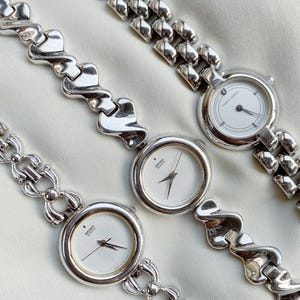 May include: Three silver-toned wristwatches with white faces and black hands. The watch bands feature unique designs, including a chain link, a wavy pattern, and a series of connected rounded rectangles. The watches are displayed on a light-colored fabric.