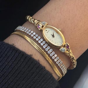 May include: A gold-tone Seiko watch with an oval face, a twisted band, and red and clear stone accents, paired with a thin gold-tone herringbone chain bracelet and a single-row clear crystal tennis bracelet on a wrist.