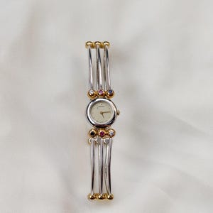 May include: A Seiko wristwatch featuring a round, off-white dial with gold hands. The band consists of three parallel silver-tone metal bars connected by gold-tone accents, with small embedded blue, red, and clear stones flanking the watch face.