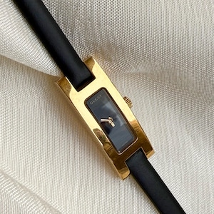 Puede incluir: Un reloj de pulsera rectangular dorado con esfera negra y correa de cuero negro. La esfera del reloj tiene la marca "Gucci" impresa. El reloj se muestra sobre una superficie texturizada de color blanco roto.