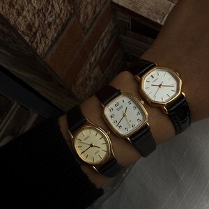May include: Three gold-toned wristwatches with black leather bands are displayed. The watches have different face shapes: round, square, and octagonal. The watch faces are white with black numerals and hands. The watches are worn together on a wrist.