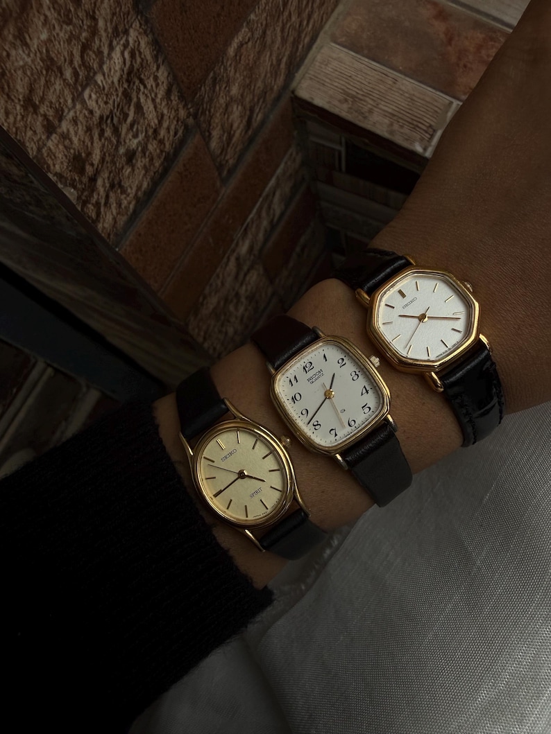 May include: Three gold-toned wristwatches with black leather bands are displayed. The watches have different face shapes: round, square, and octagonal. The watch faces are white with black numerals and hands. The watches are worn together on a wrist.