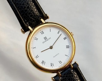 May include: A gold-toned Givenchy quartz watch with a white face and black leather band. The watch face features black Roman numerals and the Givenchy logo. The band is textured and has gold-toned hardware.