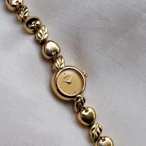 Puede incluir: Un reloj de pulsera Seiko en tono dorado con esfera redonda, manecillas e índices dorados. La correa del reloj está compuesta por una alternancia de cuentas esféricas pulidas y pequeños eslabones en forma de hoja.