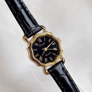 May include: A gold-toned Orient Quartz watch with a black face and Roman numeral hour markers. The watch has a black leather strap. The watch face is an octagonal shape.