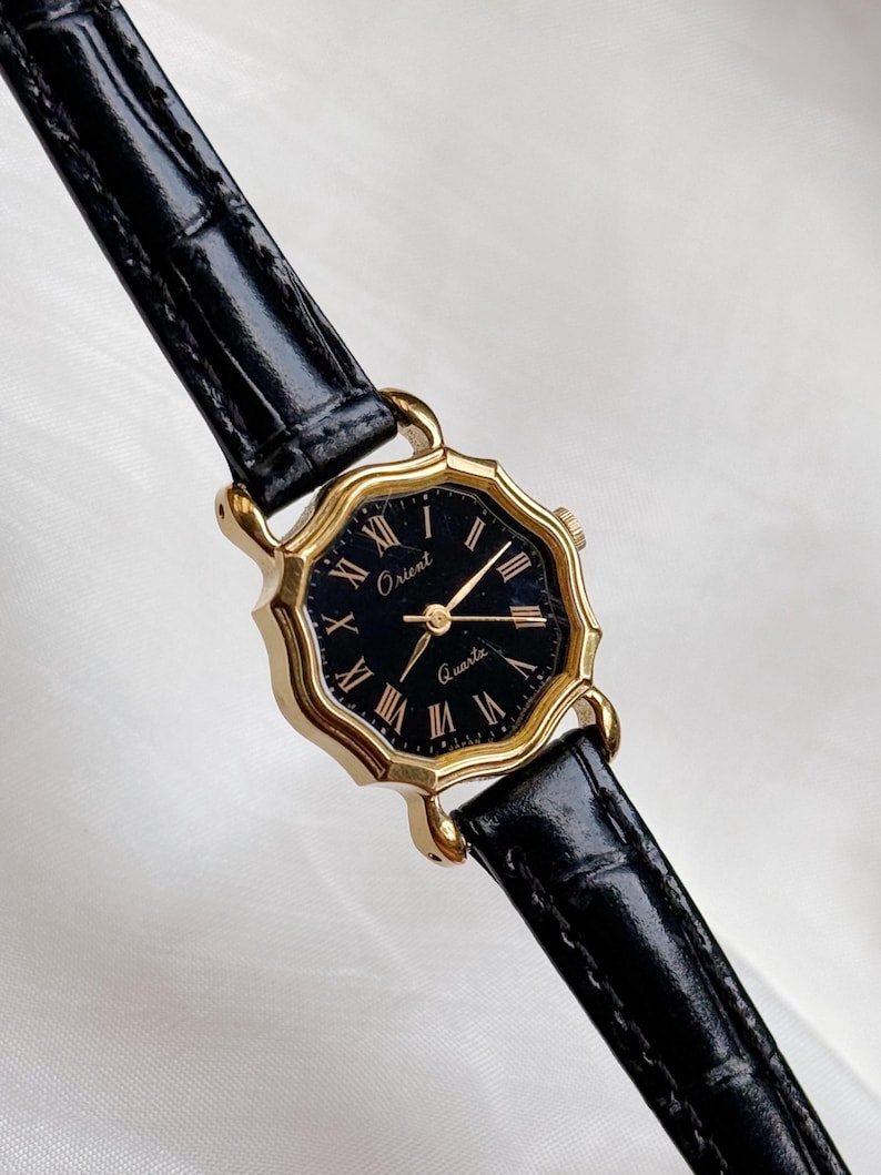 May include: A gold-toned Orient Quartz watch with a black face and Roman numeral hour markers. The watch has a black leather strap. The watch face is an octagonal shape.