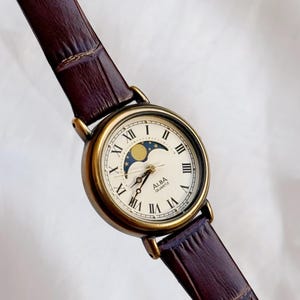 Peut inclure: Une montre-bracelet Alba Quartz vintage avec un cadran crème, un boîtier doré et un bracelet en cuir marron. Le cadran de la montre comporte des chiffres romains, un indicateur de phase lunaire et le nom de la marque. Le bracelet est marron foncé avec un motif texturé.