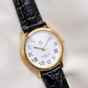 May include: A gold-tone wristwatch featuring a white dial with black Roman numeral hour markers, gold hands, and the text Givenchy Paris. The watch has a black leather strap with a textured crocodile-embossed pattern and a gold-tone crown.