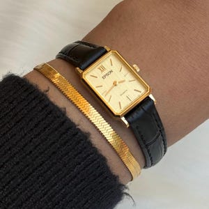 May include: A gold-toned Epson Quartz watch with a rectangular face and black leather strap. A gold herringbone bracelet is worn alongside the watch. The watch face displays Roman numerals and the Epson logo.