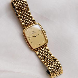 May include: A gold-tone wristwatch featuring a rectangular case with rounded corners and a textured gold dial. The watch includes a matching gold-tone metal link bracelet with a multi-row design. The Omega logo and name are printed on the dial.
