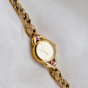 May include: A gold-toned Seiko wristwatch with a cream-colored face and a gold braided band. The watch features a small, round face with delicate hands and a moon symbol. Two small, red gemstones accent the watch face.