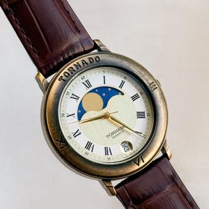 Könnte beinhalten: Eine Vintage-TORNADO-Quarzuhr mit braunem Lederarmband. Das Zifferblatt zeigt römische Ziffern, eine Mondphasenanzeige und ein Datumsfenster. Die Uhr hat eine goldfarbene Lünette und ein braunes Lederarmband.