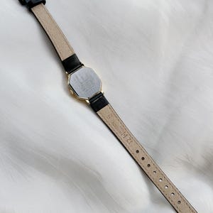 May include: A vintage gold-toned watch with a black leather band. The watch has an octagonal face and a gold buckle. The back of the watch is visible, showing the watch's details. The watch band is approximately 12 mm wide.