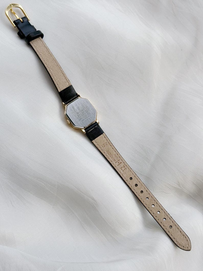 May include: A vintage gold-toned watch with a black leather band. The watch has an octagonal face and a gold buckle. The back of the watch is visible, showing the watch's details. The watch band is approximately 12 mm wide.