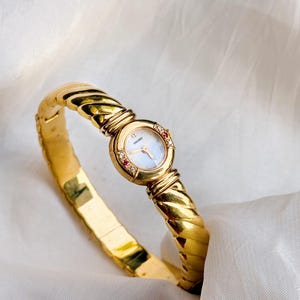 Puede incluir: Un reloj de pulsera Seiko dorado con una pulsera trenzada. La esfera del reloj es redonda con un efecto nacarado claro. Pequeñas piedras rojas y transparentes adornan la esfera. El reloj se muestra sobre una superficie blanca.