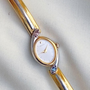 Puede incluir: Un reloj de pulsera Seiko dorado y plateado con esfera blanca y agujas doradas. El reloj tiene un bisel y una correa dorados con detalles plateados. La correa está adornada con dos piedras preciosas, una rosa y otra azul.