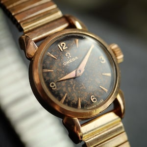 May include: Close-up of a vintage Omega wristwatch with a gold-toned case and band. The watch face is dark with gold numerals and hands. The band is a flexible, expanding style.