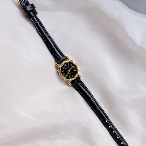 May include: A black wristwatch with a gold octagonal face and black leather strap. The watch has Roman numeral hour markers and a gold buckle. The strap is approximately 1.3 cm wide.